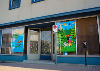 Two large illustrated posters hanging in an empty storefront window by Jennifer Ilett. The illustrations showcase people canoeing and hiking.