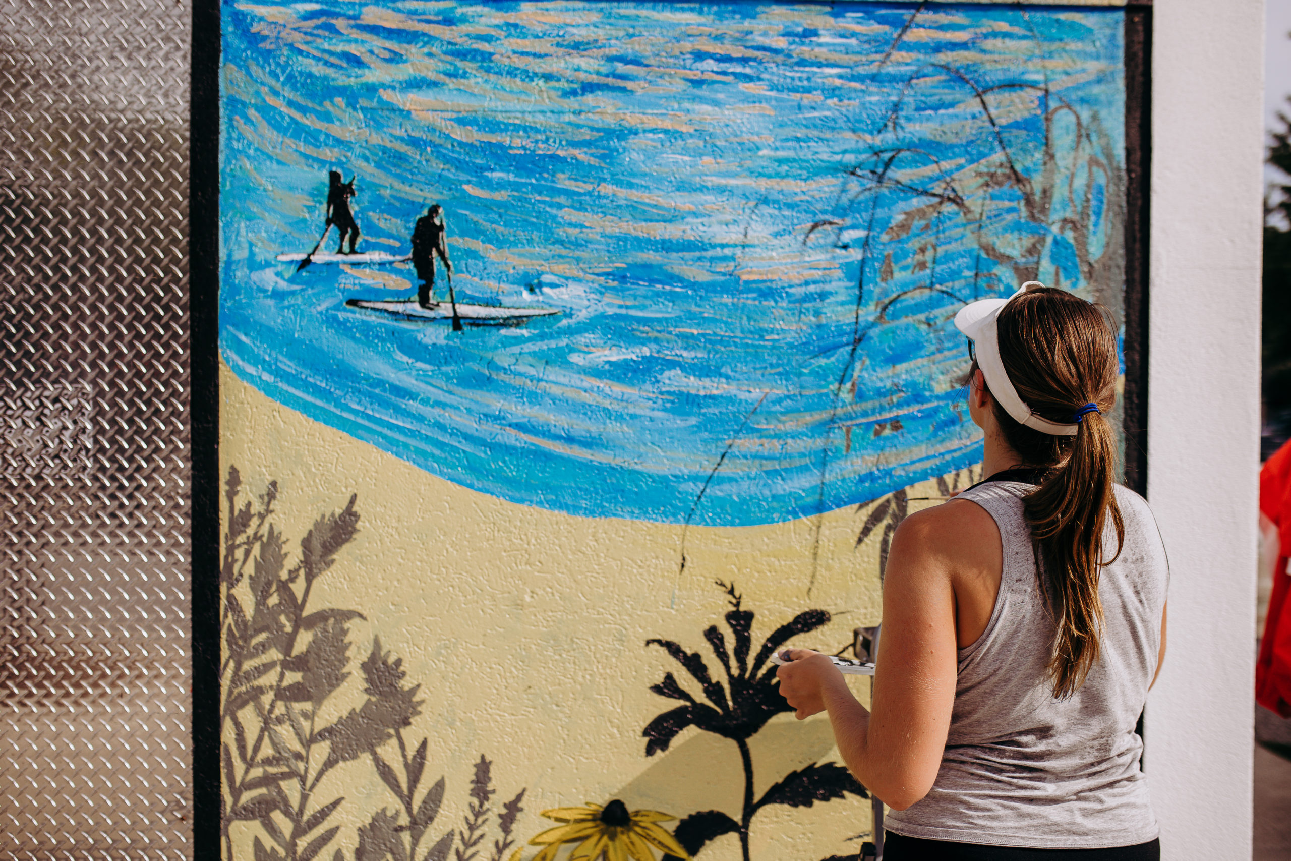 Jocelyn Kearns painting a large outdoor mural depicting soft waters with people paddle boarding.