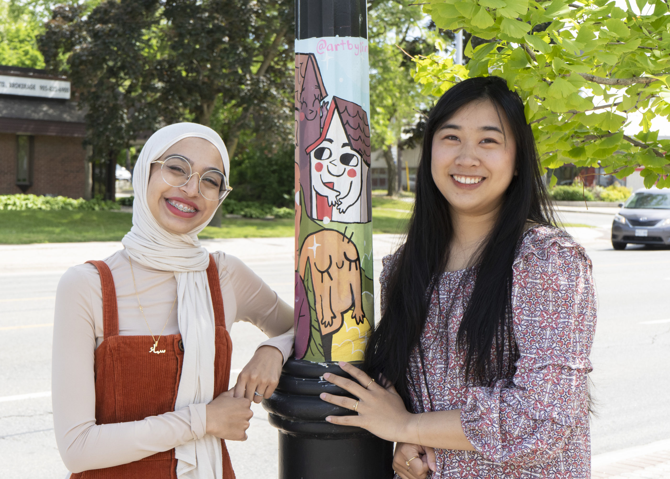 Artists Sima Naseem and Yen Linh Thai posing beside a painted public art mural on a street pole in Clarkson Village, Mississauga