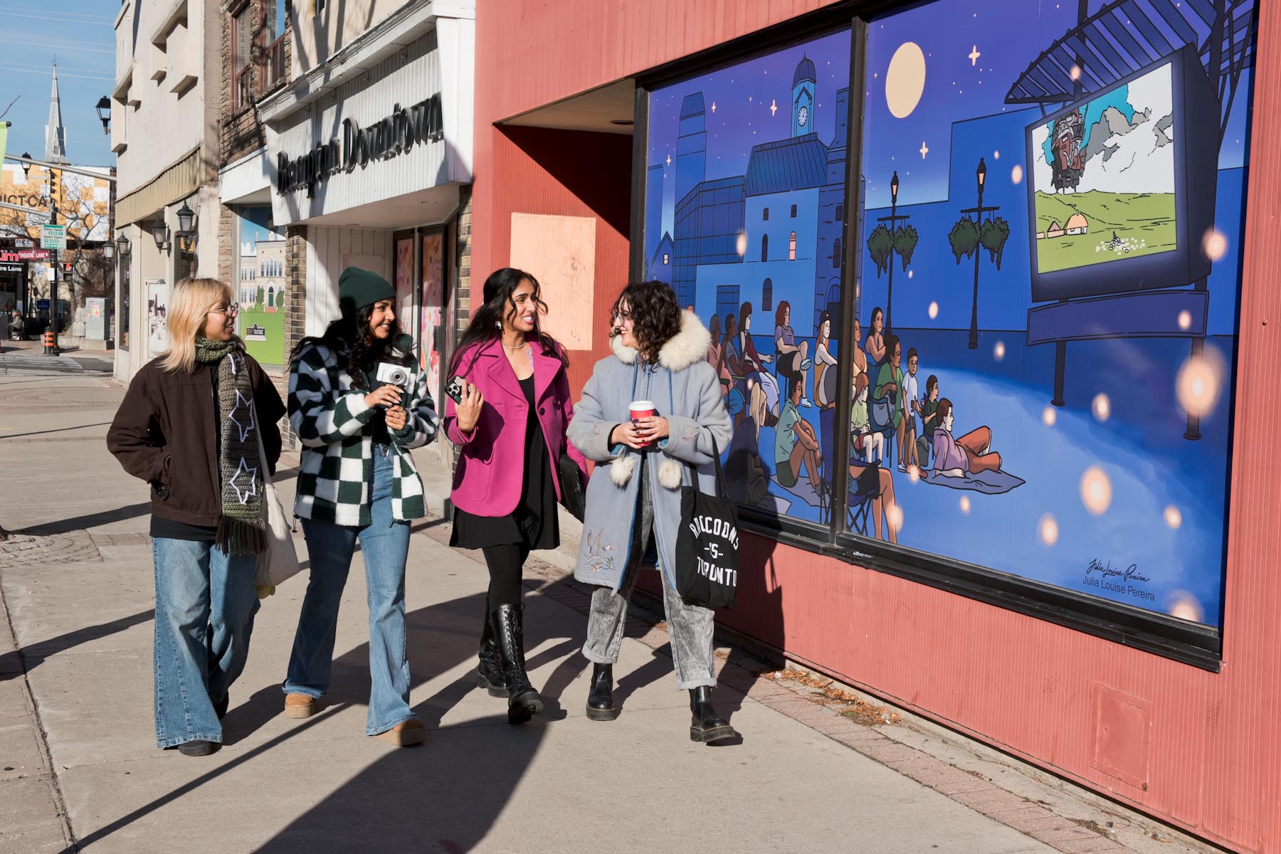 Photo of 4 artists (left to right: Neha Ray, Saphera Peters, Sabbie Narwal, Julia Louise Pereira) walking and talking along Main Street North in Brampton, where artwork by Saphera and Julia are installed on the storefronts.