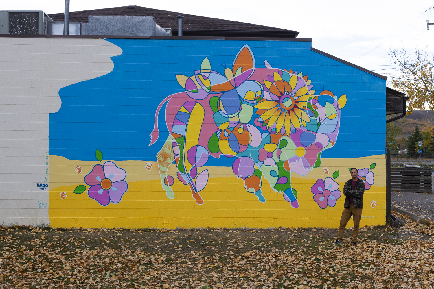 Photo of Bruno Canadien’s mural “Source of Life” it is on a brick building, Bruno is standing in front of it, looking at the camera It features geometric shapes and motifs that form a buffalo - there are colours of pink, green, yellow, and purple on a blue background. Part of the overcast sky is showing on the top right corner.