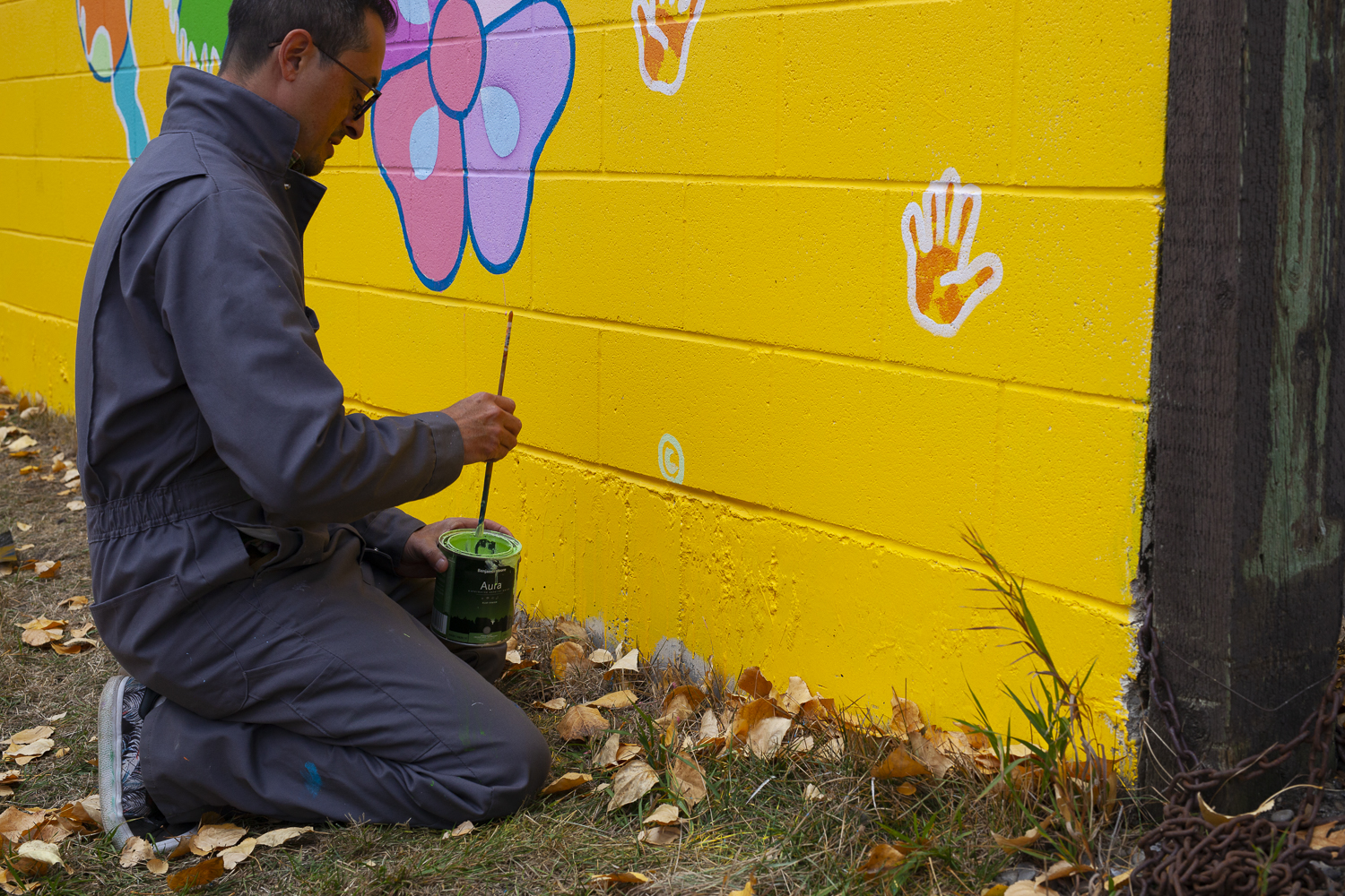 Photo of Bruno Canadien dipping a paintbrush in green paint, he is kneeling on some grass with leaves, about to paint his mural. The mural has colours of pink, purple, blue and orange on a yellow background.