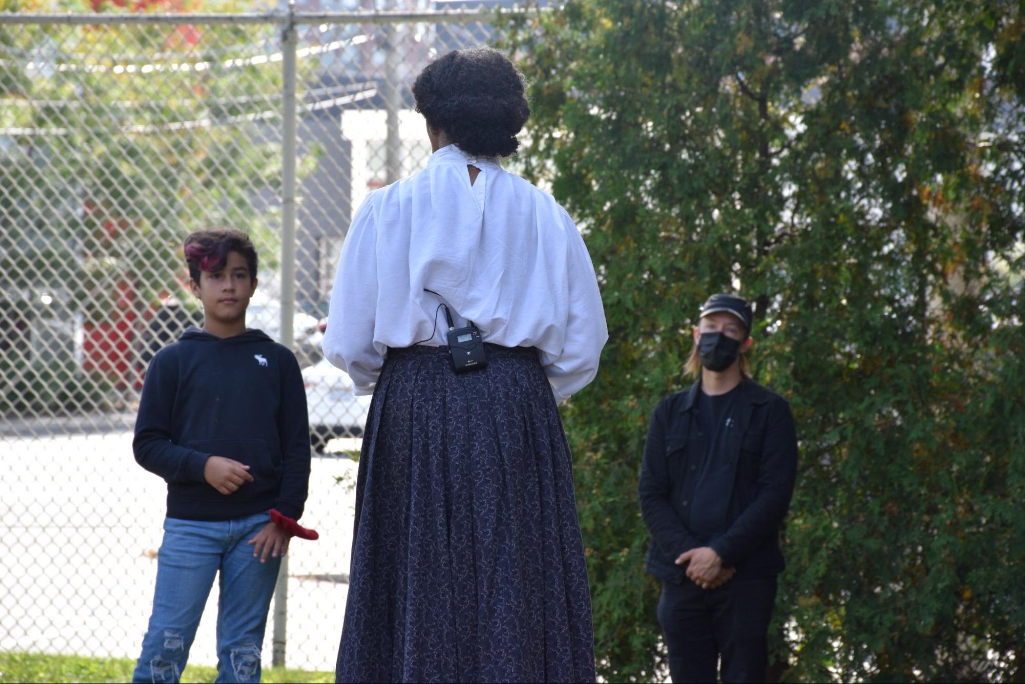 Photo of Charmaine Lurch turned around, talking to two people, she is wearing a dark skirt, white shirt. There is a wire fence and greenery in the background.