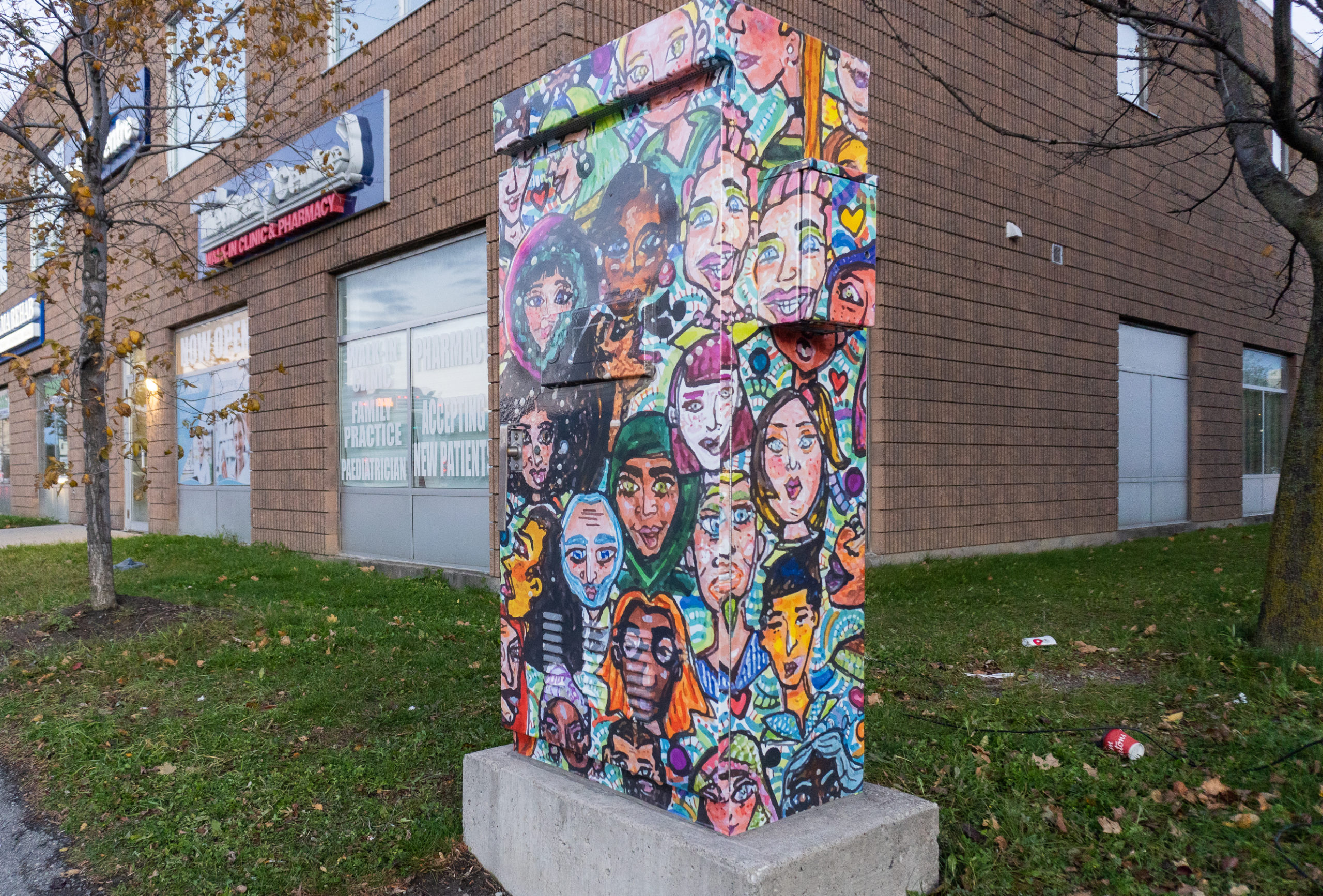Photo of a traffic box in Brampton that features the artwork of Annmarie Claudette. The art shows a collage of colourful portraits with bold patterning. The box is sitting on a grassy area next to the building of a pharmacy and trees.