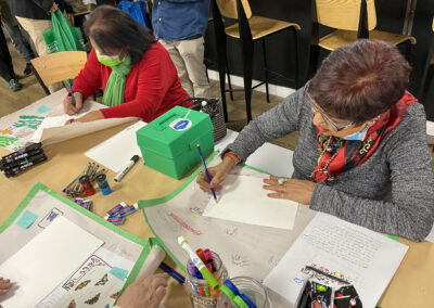 Image of two workshop participants drawing with arts supplies on a table covered by kraft paper.