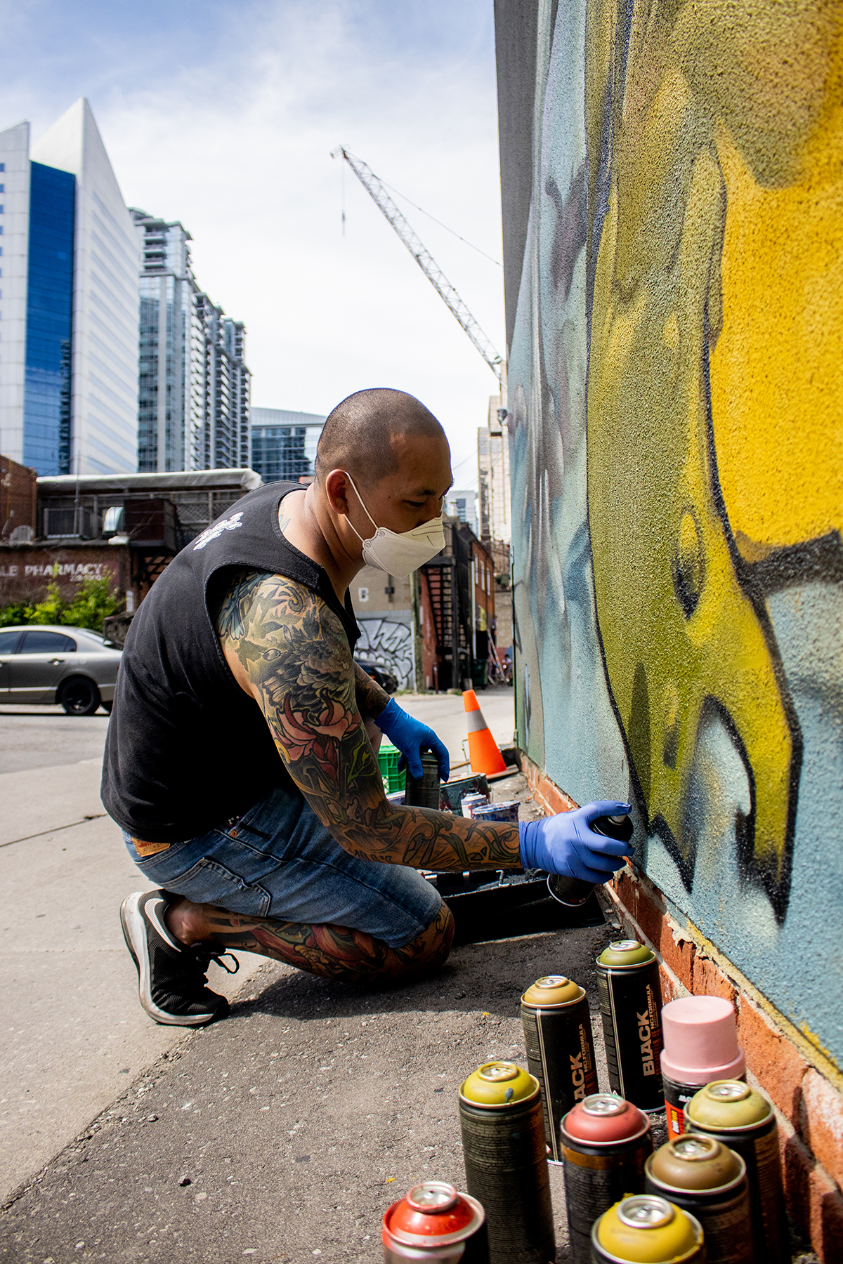 Photograph of artist Eugene Lee painting his mural on a building, it features a large frog/toad that is olive green and yellow in colour. There is also a red snail painting to the right side of the image.