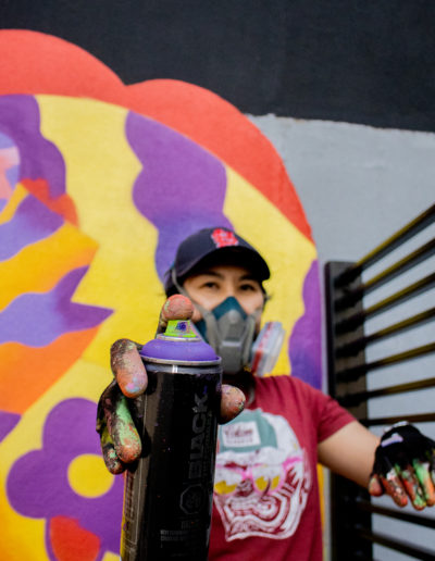 Photo of Jieun June Kim holding a purple spray paint can to the camera - background is her mural with colours of red, purple and yellow