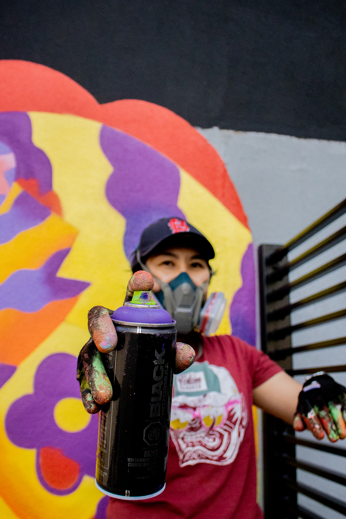 Photo of Jieun June Kim holding a purple spray paint can to the camera - background is her mural with colours of red, purple and yellow