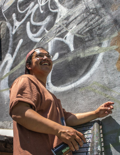 Photograph of artist Luvs, he is looking upwards and smiling, wearing a brown shirt. He is in front of a wall that is grey and has some black and white graffiti art.