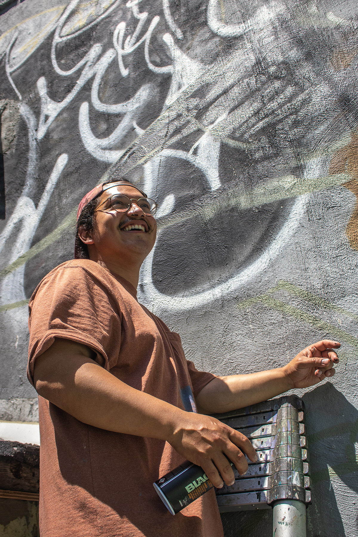 Photograph of artist Luvs, he is looking upwards and smiling, wearing a brown shirt. He is in front of a wall that is grey and has some black and white graffiti art.