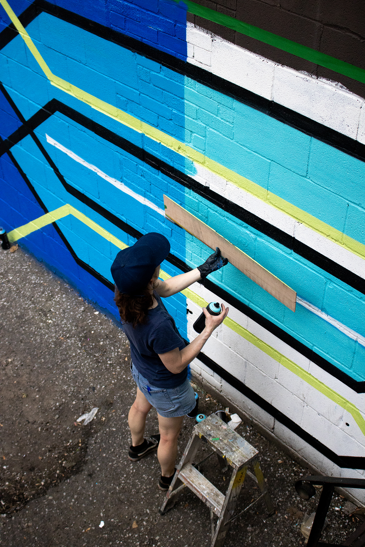 Photograph of artist Erika James painting her mural that features geometric lines and shapes in blue, teal, green and white. She is wearing a dark-colored t-shirt and baseball cap.