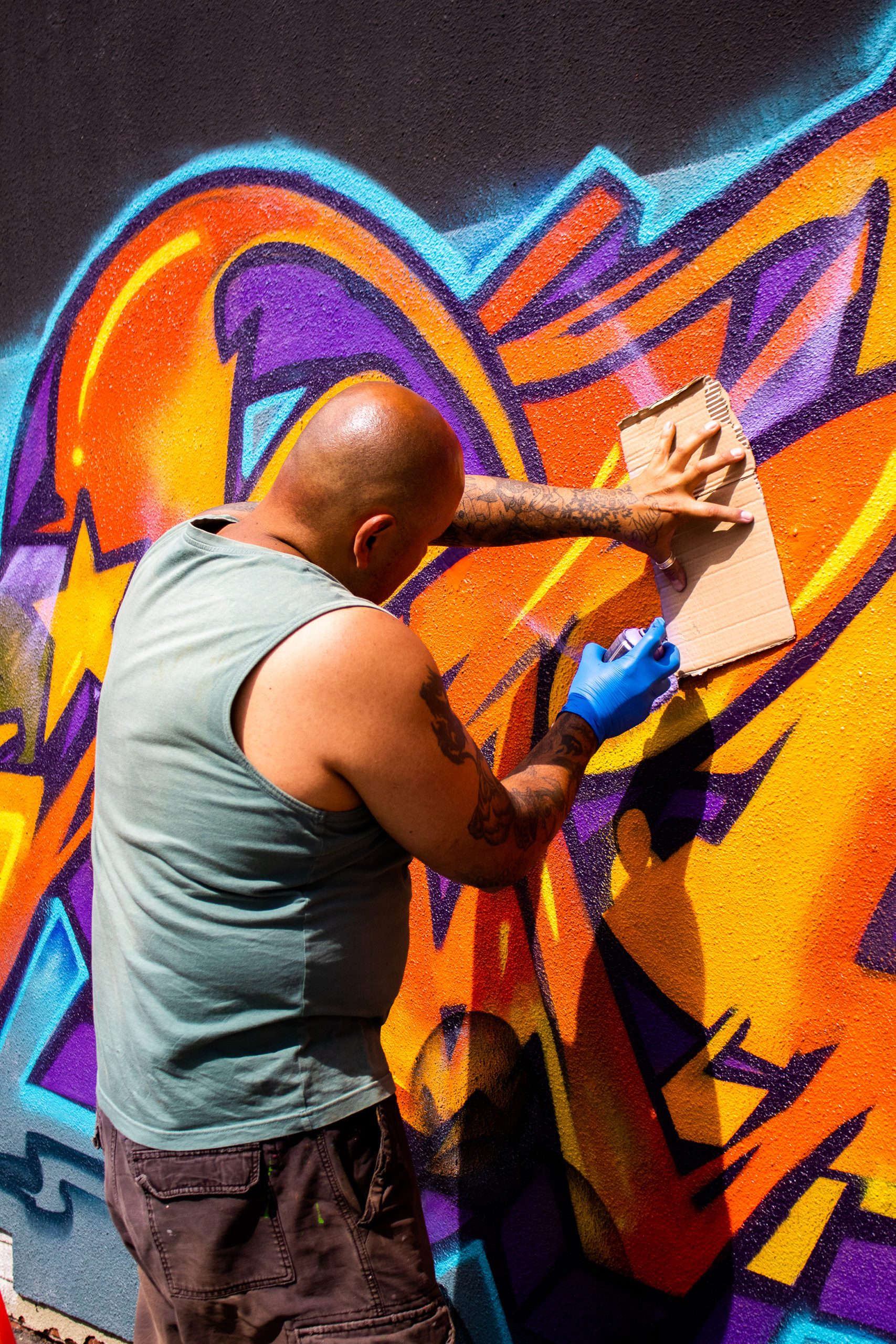 Photograph of artist Angel Cruz painting his mural on a wall. There are bright colours of orange, red, purple and blue present.