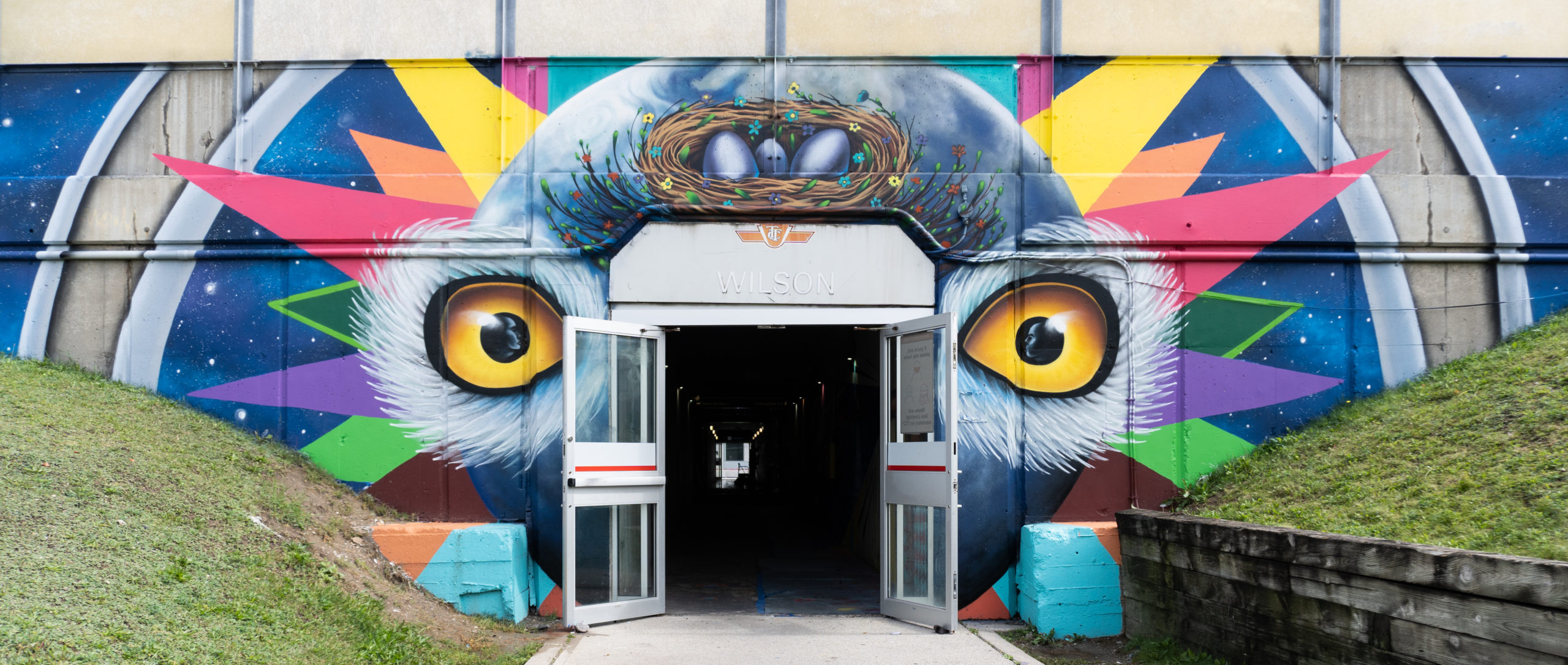 Photograph of mural by Shalak Attack at Wilson TTC Station entrance. The doors are wide open leading to the subway, the mural is of an abstracted bird with vibrant gold eyes and a blue “head” that is resemblant of a globe. There is a nest with eggs above it’s head and colourful geometric shapes radiating around the focus.