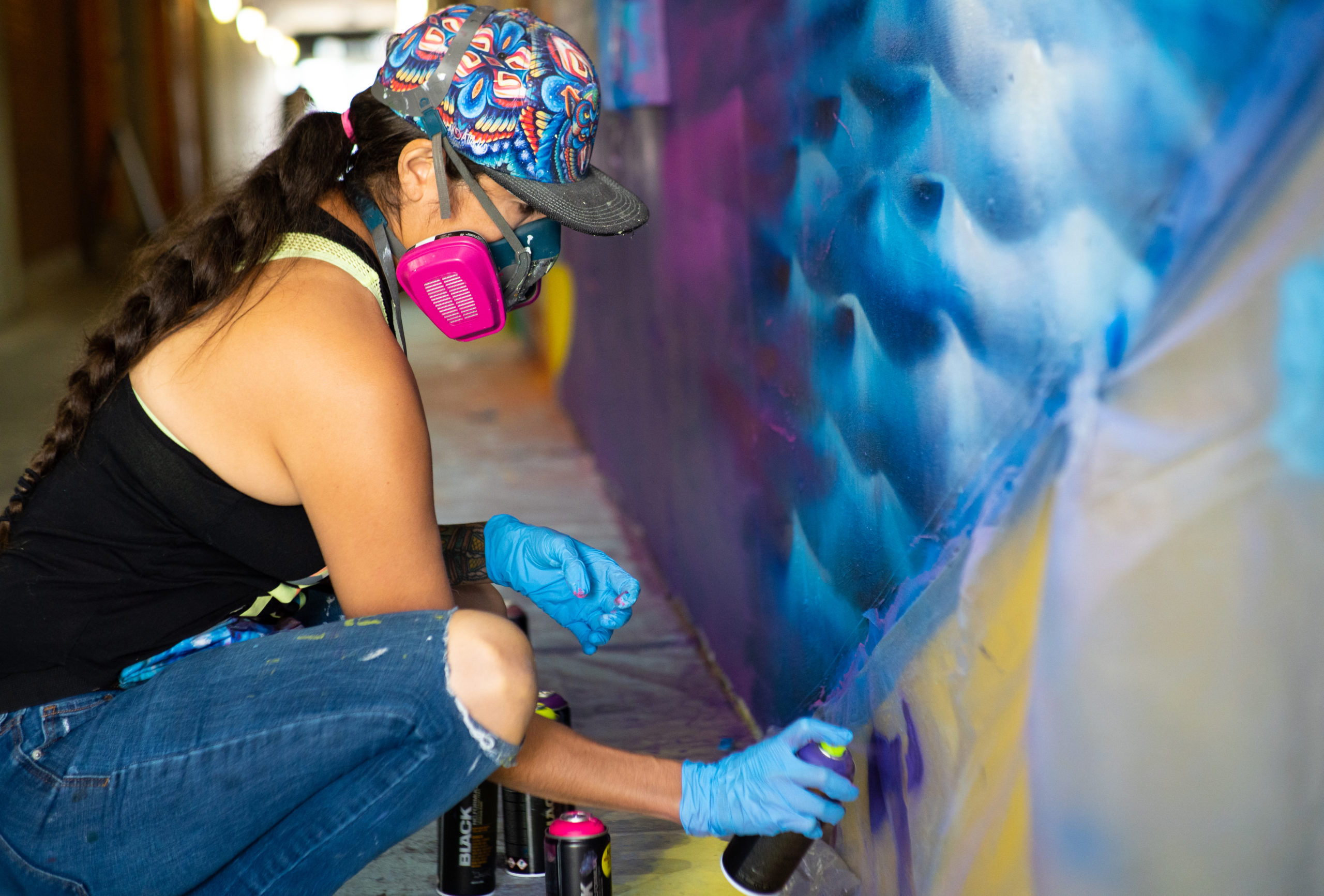 Photo of artist Shalak Attack inside Wilson TTC Station spray painting a portion of her mural with some blue waves. Shalak is wearing a black tank top, jeans, patterned hat and latex gloves.