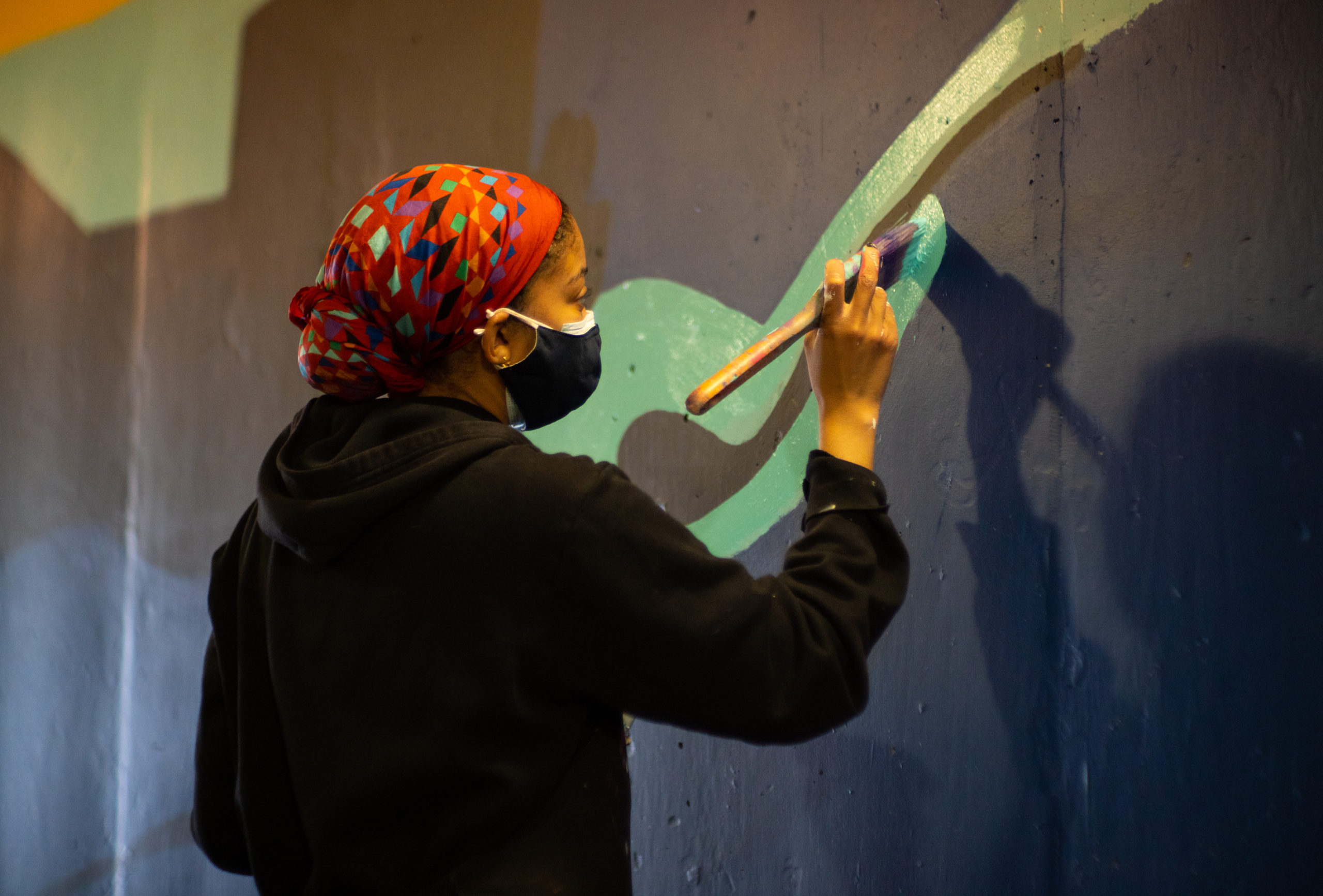 Photograph of art assistant Edan Maxam painting a mural in the entrance of Wilson TTC Station. Edan is wearing a mask, her hair is wrapped with a patterned fabric and she is wearing a black hoodie. The mural has colours of navy blue and green.