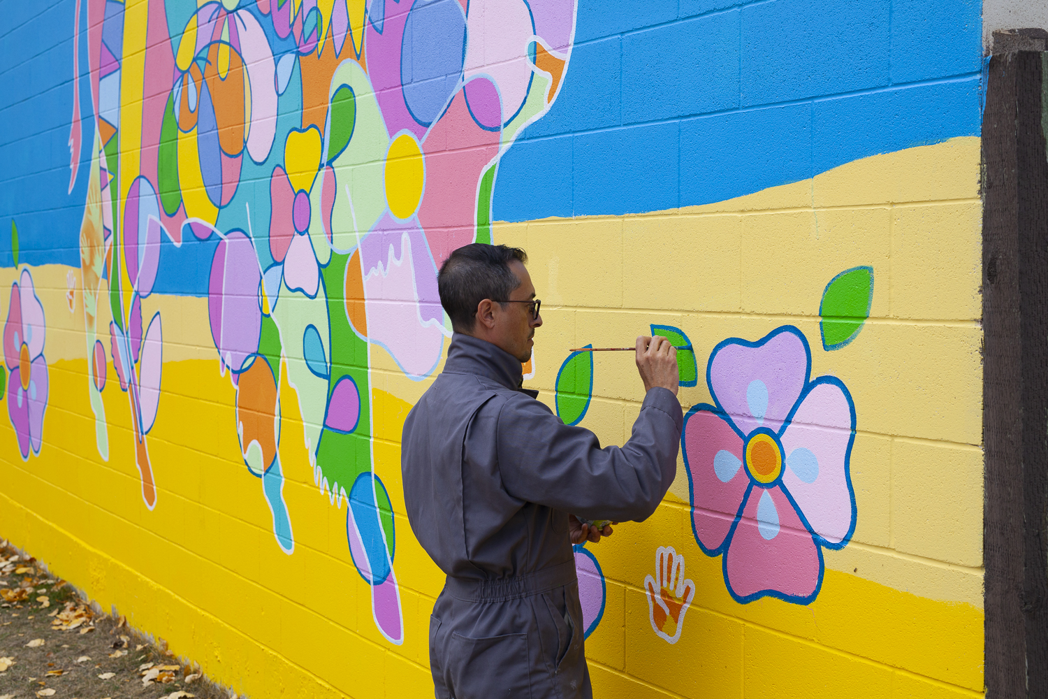 Photo of Bruno Canadien painting a leaf detail on his mural “Source of Life”. The mural features geometric shapes and motifs that form a buffalo - there are colours of pink, green, yellow, and purple on a blue & yellow background.