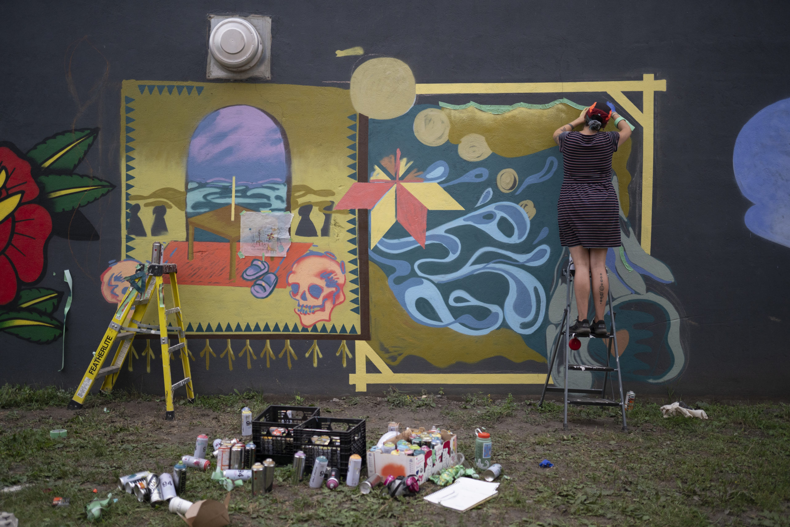 Photo of artist Shelby Gagnon on a step ladders and painting her mural, there are spray cans and supplies scattered around scene. The mural has colours of yellow ochre, purple, blue and red.