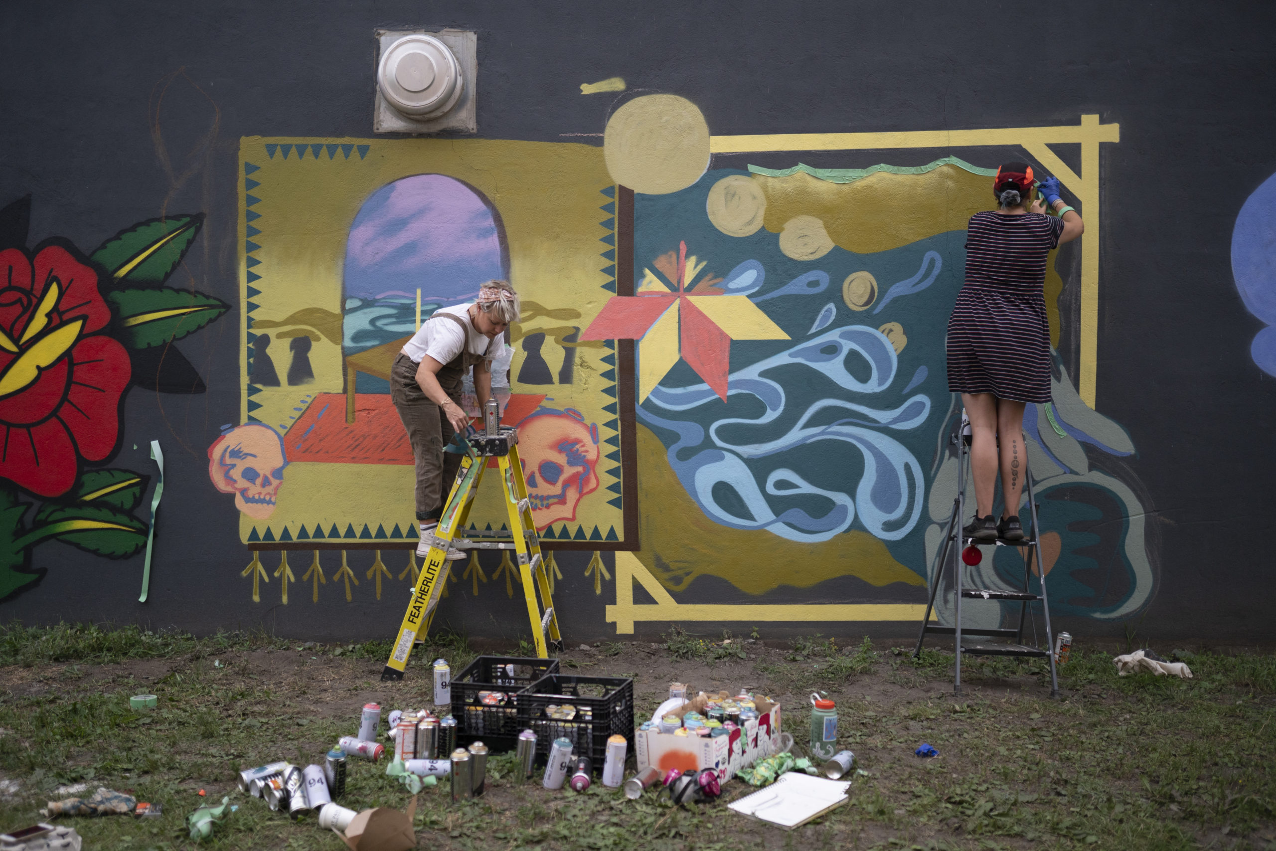 Photo of artist Shelby Gagnon and her mentor Lora Northway painting their mural. Both are on step ladders, there are spray cans and supplies scattered around scene. The mural has colours of yellow ochre, purple, blue and red.