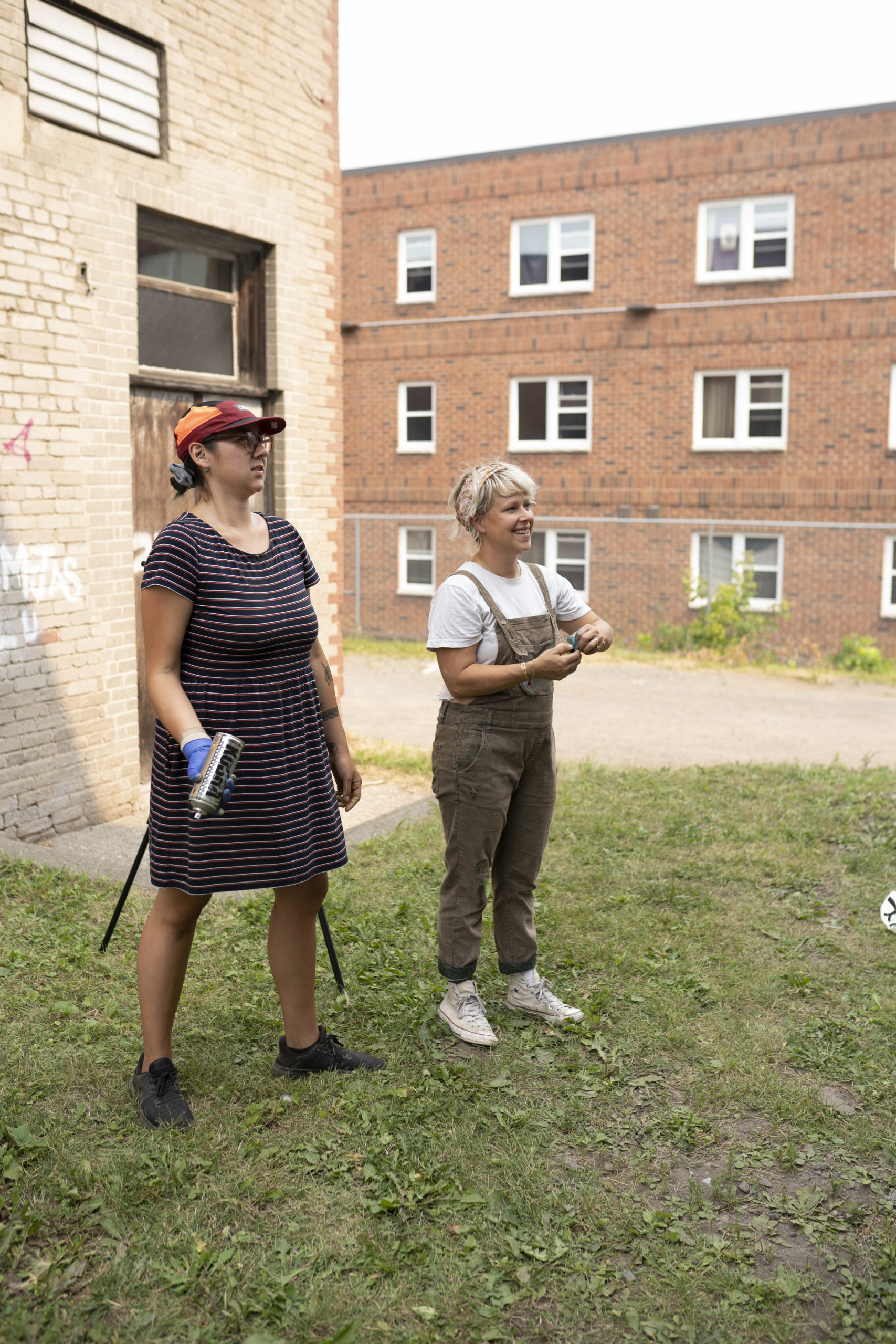 Photo of artist Shelby Gagnon and her artist mentor Lora Northway, they are smiling and looking at their mural. They are standing on green grass and two brick buildings are in the background.