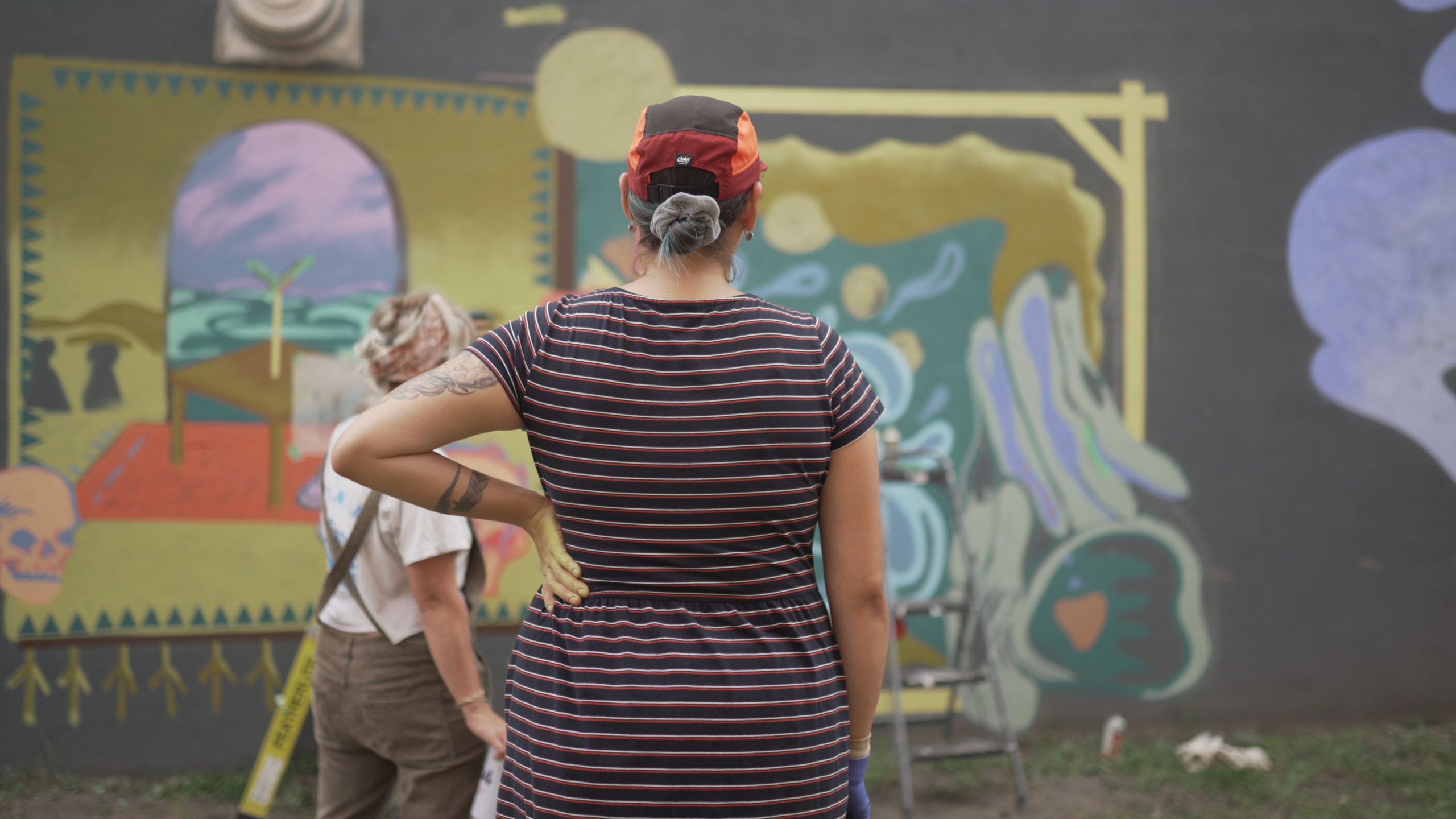 CreateSpace artist Shelby mentor wearing a striped dress and orange hat, look at her mural. Her left hand is on her hip and the other is straight, and her hair is tied into a bun. Her mentor Lora Northway is visible on the left and the mural has muted colours of yellow, teal, purple and red.