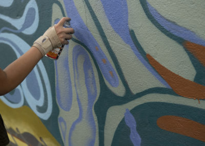 Hand spray-painting a mural with muted organic shapes with colours of yellow, purple, red and teal.