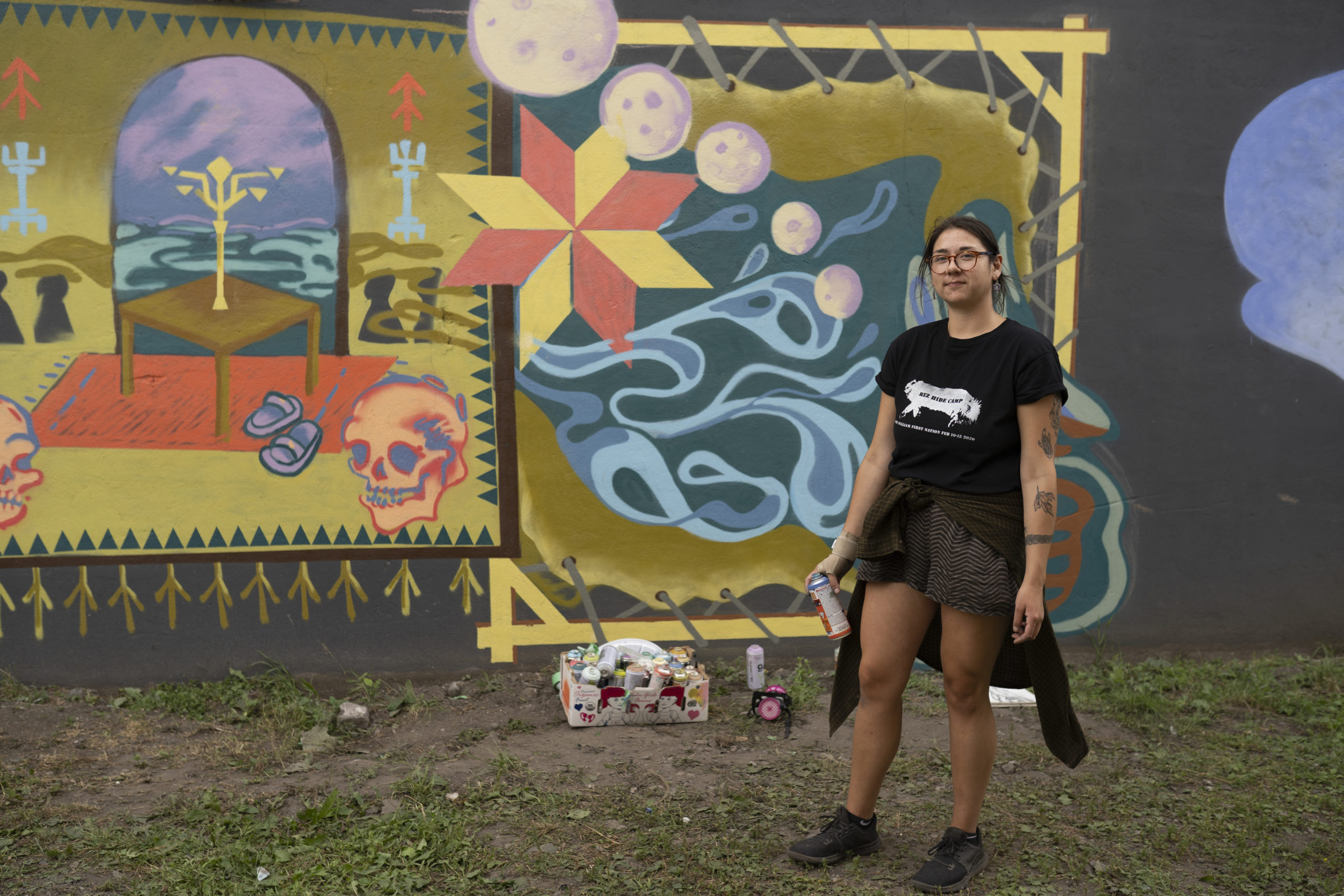 Artist Shelby Gagnon posing in front of her first mural that has both Anishinaabe and Finnish designs. It has colours of yellow ochre, blue, purple and red. Shelby is wearing an all black outfit and parts of the grass is showing.