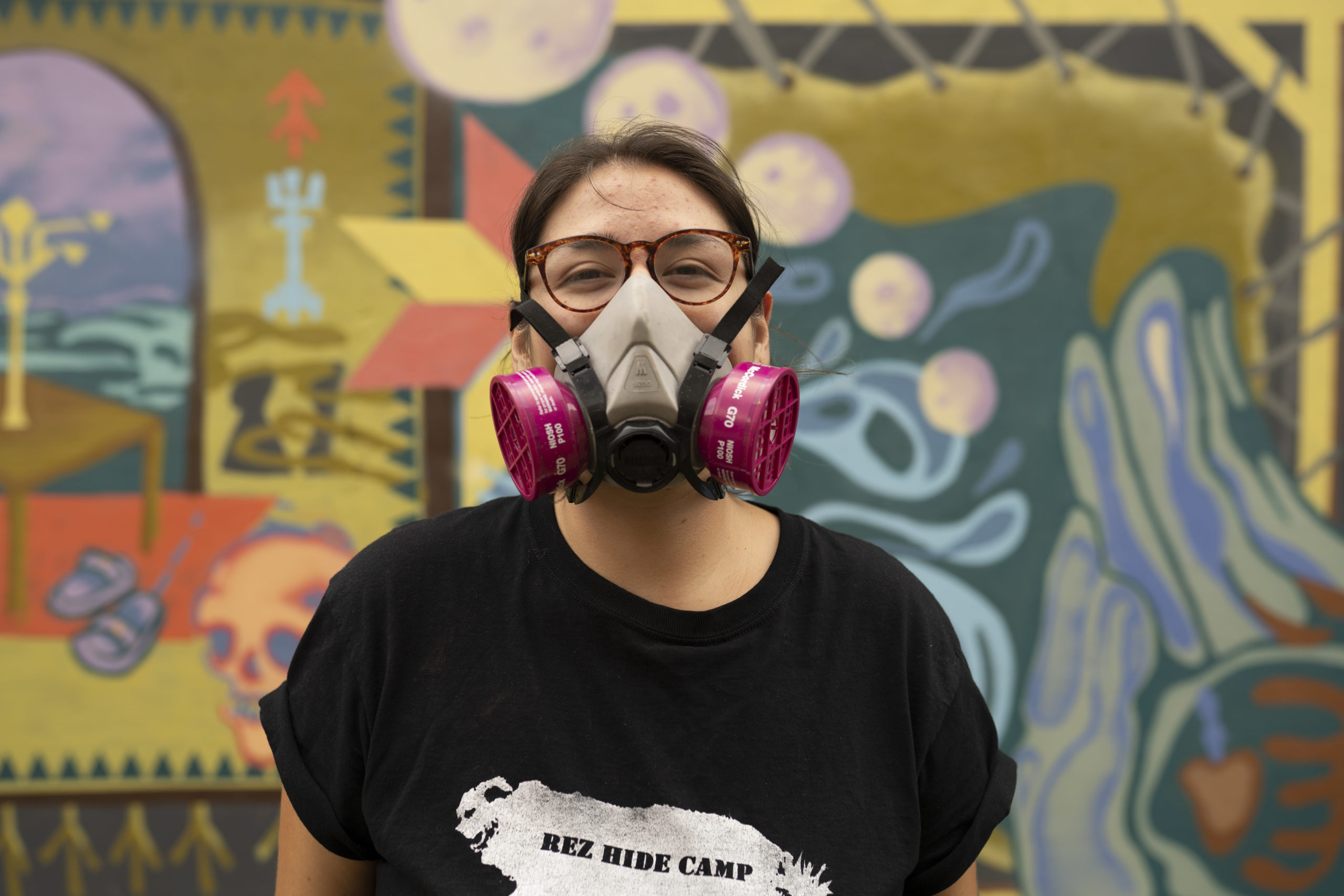 Artist Shelby Gagnon looking at camera, smiling with a spray paint mask on and a graphic tee that reads "REZ HIDE CAMP". She is standing in front of her first mural with colours of yellow ochre, purple, teal and orange.
