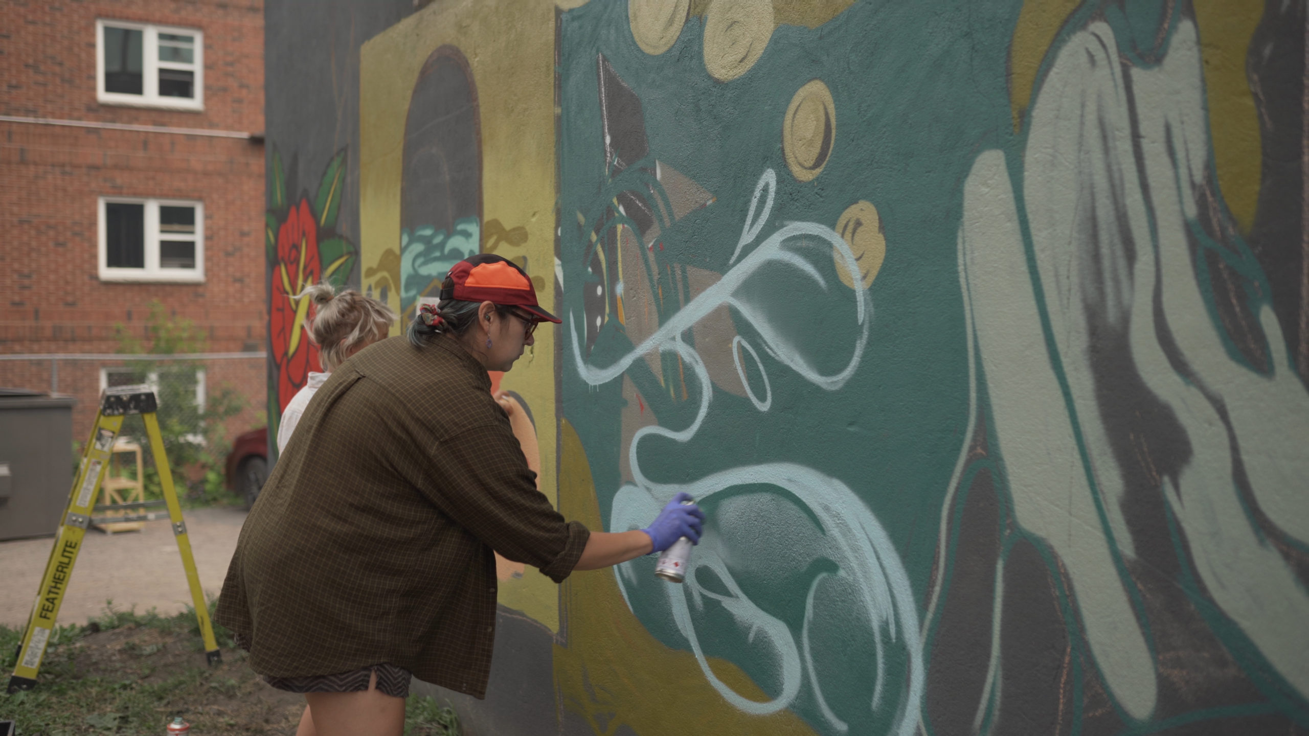 CreateSpace artist Shelby Gagnon spray painting an organic shape for her mural on Haven Hostel. Shelby is wearing a blue shirt, orange hat and purple latex gloves. The mural has muted colours and compliments he neutrals of the location. There is a yellow in the left side.
