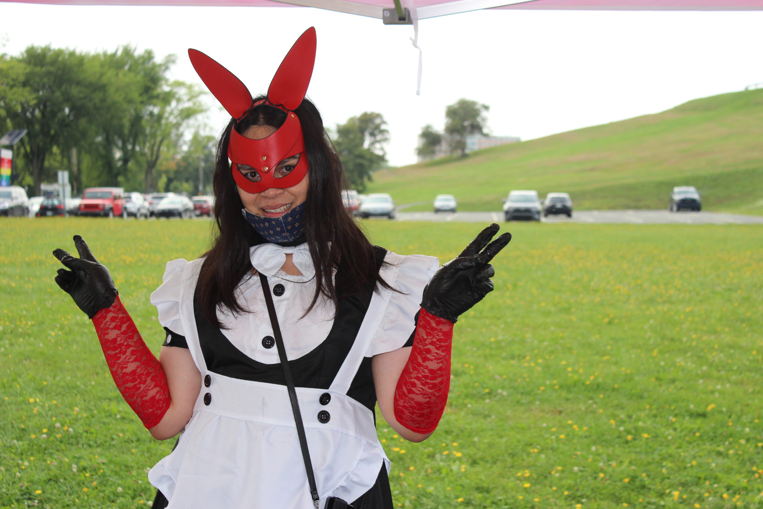Photo of Excel Gray looking at the camera, smiling and holding up their hands with peace signs. They are wearing a black & white dress, black and red gloves, a face mask, red face covering with bunny ears. They are standing in a green field with a parking lot in the distance.
