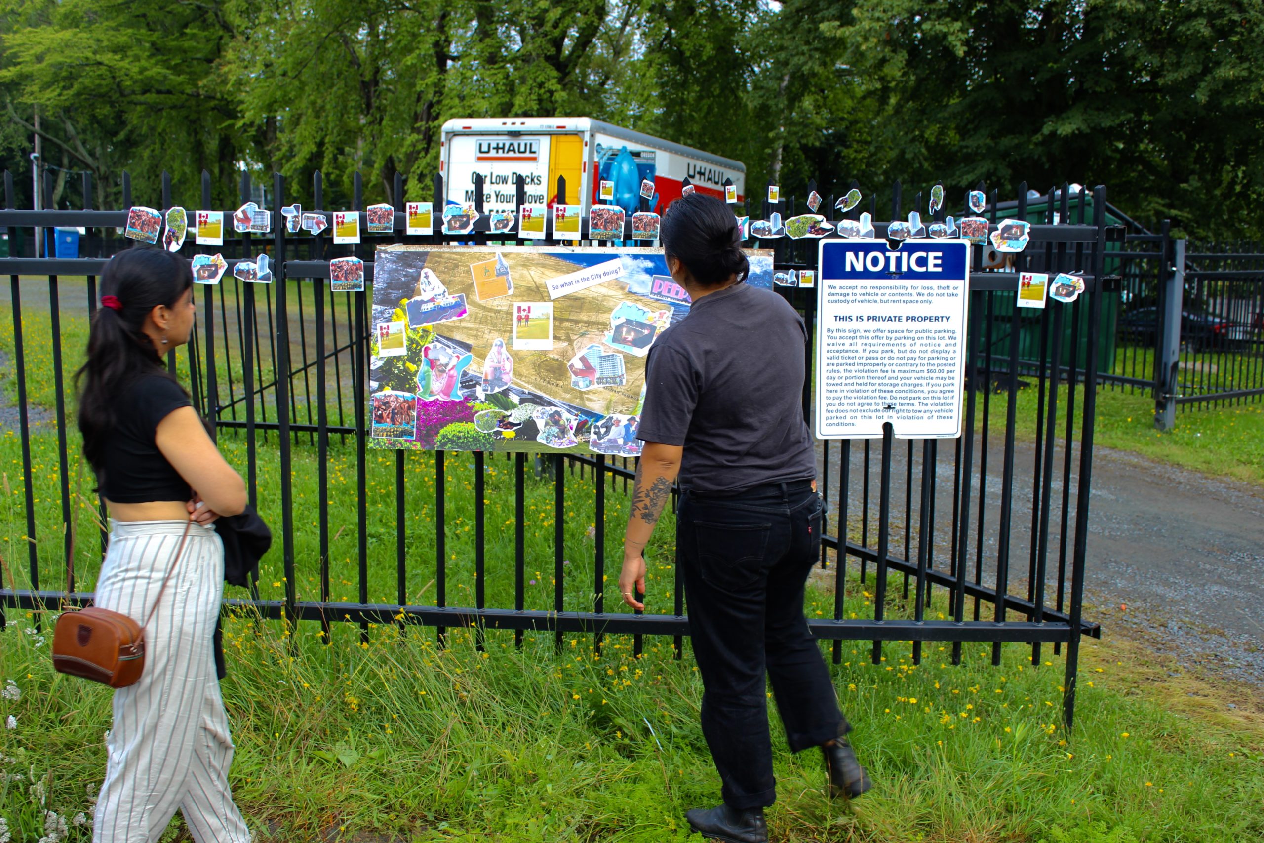 Photo of two people looking at an art installation by Arjun Lal and collaborators, it features small cut photos and collages along a black metal fence, it also has one large poster attached with collaged imagery of various colours. The fence is on a grassy area of a part, with a pathway and trees in the background.