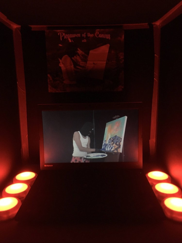 Photo of a laptop in a dark box with warm lights illuminating it slightly, the laptop is playing a video a person in a white tank top, ski mask and gloves painting a canvas on an easel.