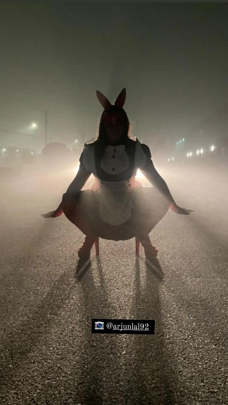 Photo of Excel Garay at night wearing a mask with bunny ears, black & white dress, heeled shoes and gloves. There is a car with its headlights on, creating a silhouette of Excel.