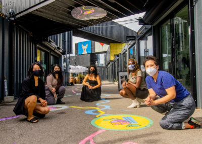 Representatives from STEPS and Stackt Market kneeling on the ground with artist Amanda Lederle, beside their finished ground mural. Its colourful and has round badges with organic lines connected them. It is located at Stackt Market with multiple black converted shipping containers on the premises. Everyone is wearing masks.
