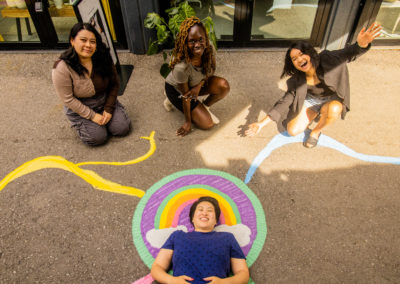 Three STEPS representatives kneeling on the ground beside Amanda Lederle and their ground mural "Life Badges" at Stackt Market. Amanda is in the center of the image, laying on the ground with their mural of a rainbow badge. Parts of the shipping container storefronts are visible.