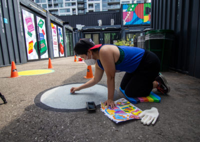 Photo of Amanda Lederle sketching out a design on their ground mural, holding onto chalk and a sketch of the design. There are pylons scattered around Stackt Market, with black converted shipping containers with signage throughout.