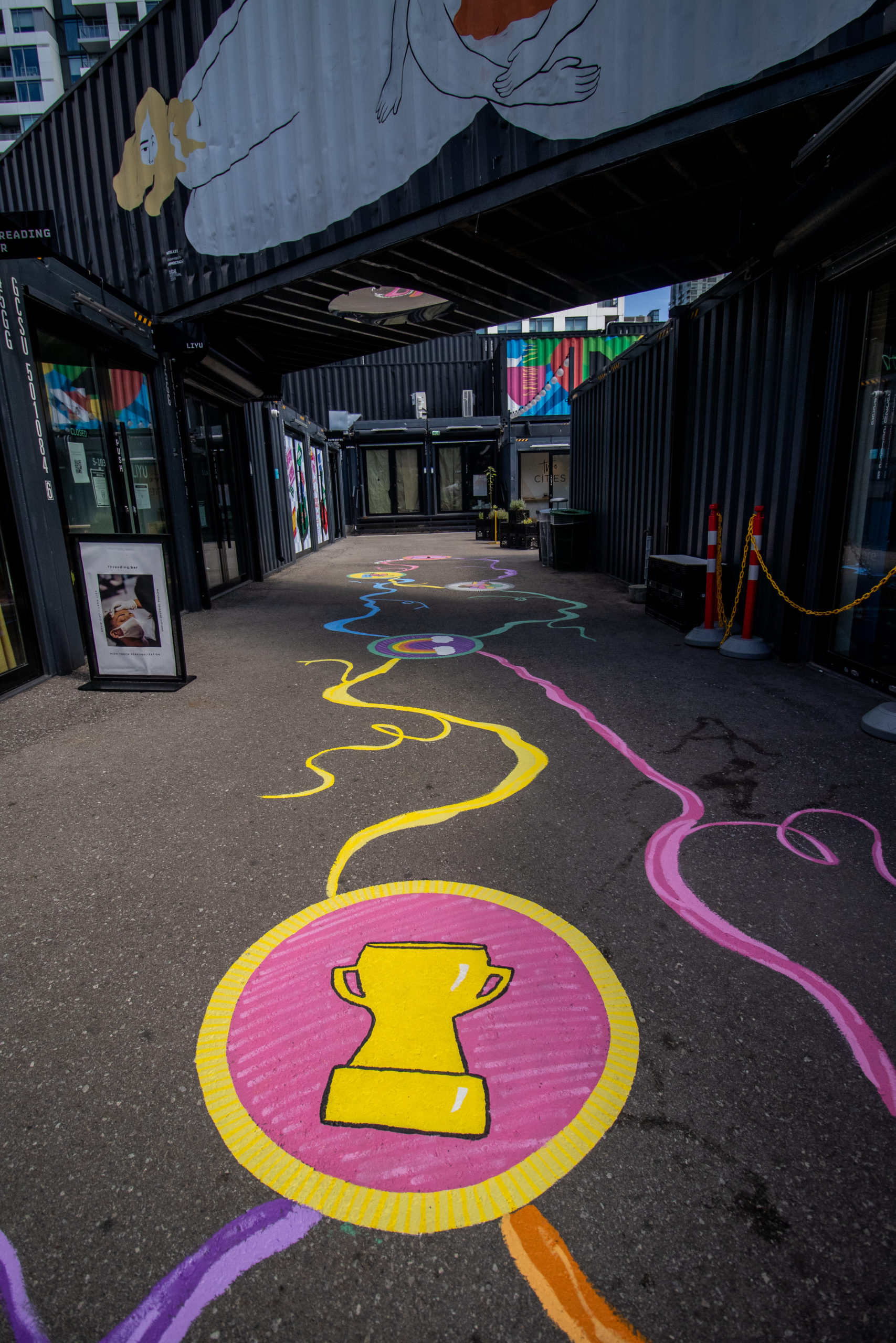 Vertical photo of a ground mural by Amanda Lederle that has "badges" with organic lines attaching them, it is colourful and bright. Located at Stackt Market in Toronto, with many converted black shippin containers, one on top with a mural by Ness Lee and condos in the back.