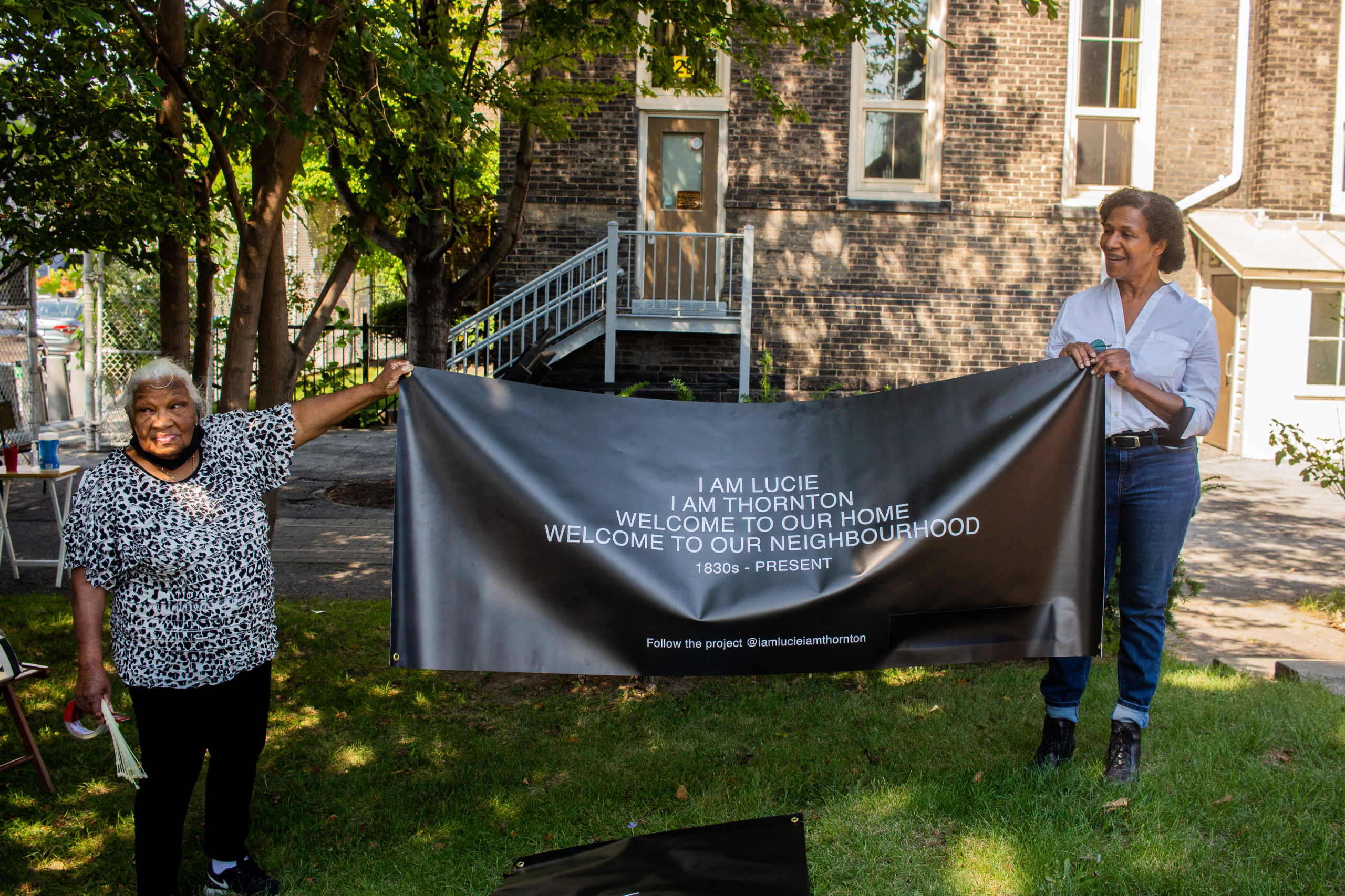 Photo of Charmaine Lurch and a woman holding up a black and white banner for her project "I Am Lucie, I Am Thornton". They are on the site of Lucie and Thornton's historical home, and there is grass and trees surrounding it.