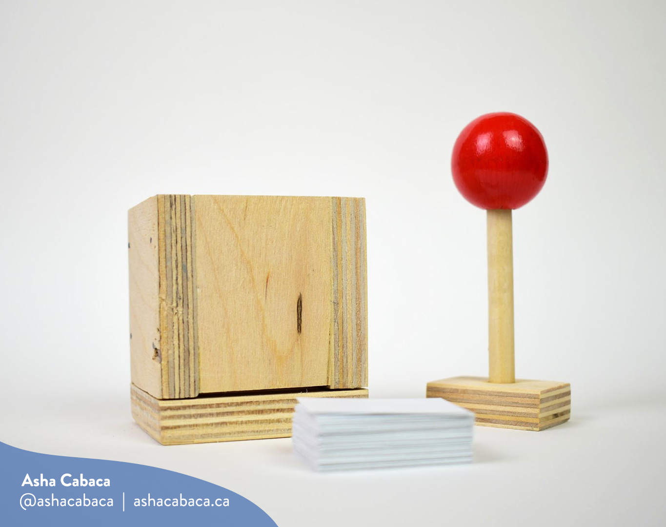Horizontal photo of artwork by Asha Cabaca, it features a wooden box & a stick with a red handle used to shape mud into a brick, it lays on a white surface with a stack of paper. There is a watermark in blue that has Asha’s name, Instagram handle (@ashacabaca) and website (ashacabaca.ca).