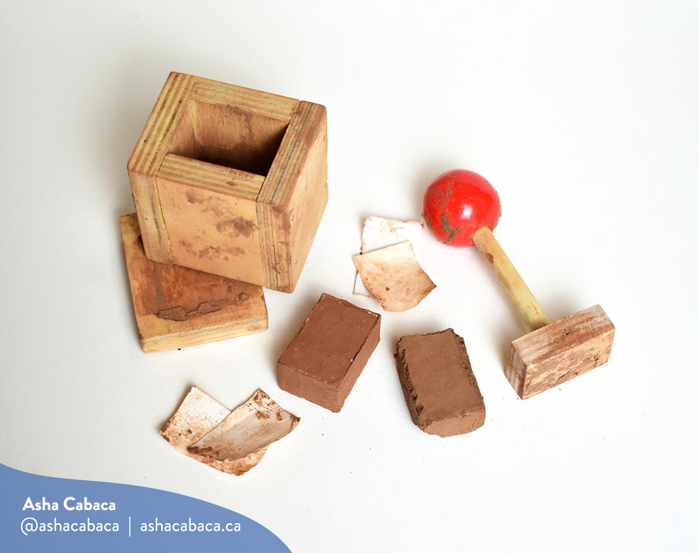 Horizontal photo of artwork by Asha Cabaca, it features a wooden box & a stick with a red handle used to shape mud into a brick, it lays on a white surface. There is a watermark in blue that has Asha’s name, Instagram handle (@ashacabaca) and website (ashacabaca.ca).