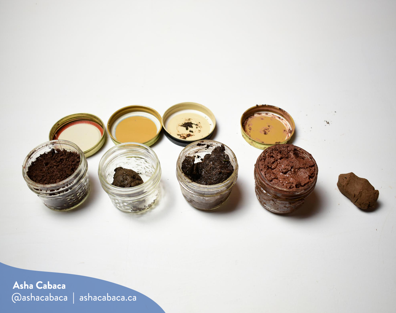 Horizontal photo of artwork by Asha Cabaca, it features four kinds of mud in four glass mason jars on a white surface. There is a watermark in blue that has Asha’s name, Instagram handle (@ashacabaca) and website (ashacabaca.ca).