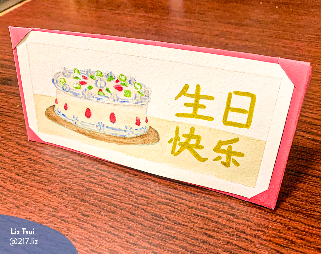 Horizontal photo of a card illustration by Liz Tsui, showcases a Chinese fruit cake on a table with Chinese characters. The card is standing up on a redwood table. The photo has a watermark with Liz’ name and Instagram handle (@217.liz).
