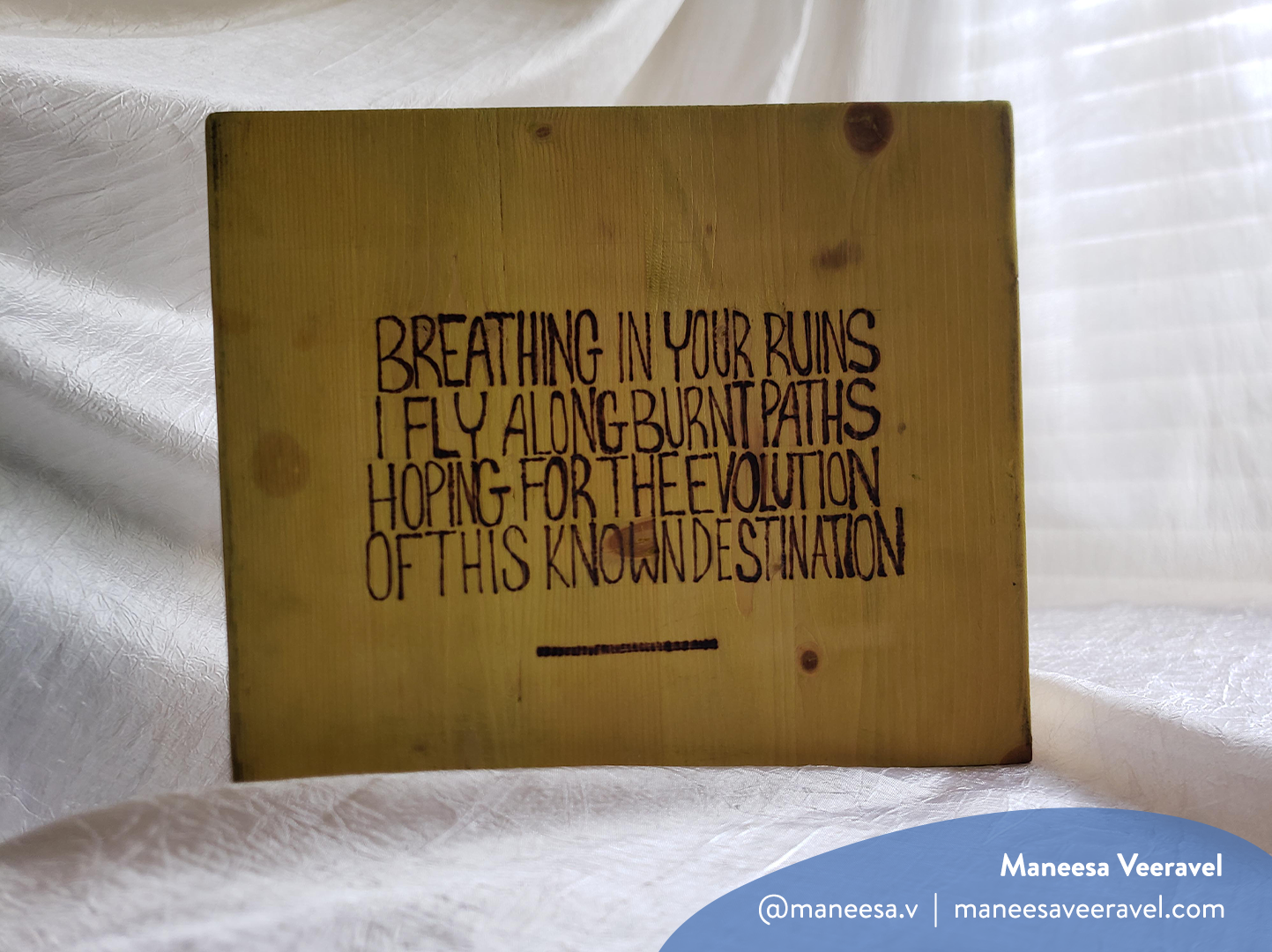 Horizontal photo of wood-burned artwork on a white fabric background by Maneesa Veeravel. It features hand-lettered text reading: “Breathing in your ruins, I fly along burnt paths, hoping for the evolution of this known destination”. The image has a watermark with Maneesa’s name, Instagram handle (@maneesa.v) and website (maneesaveeravel.com)