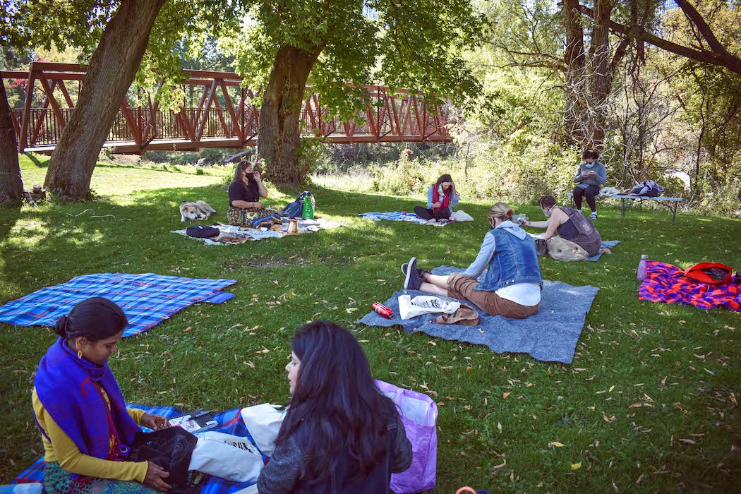 Photograph of a communal art gathering in a park, where the participants are engaging in a beading workshop. In the background are trees and a red bridge.
