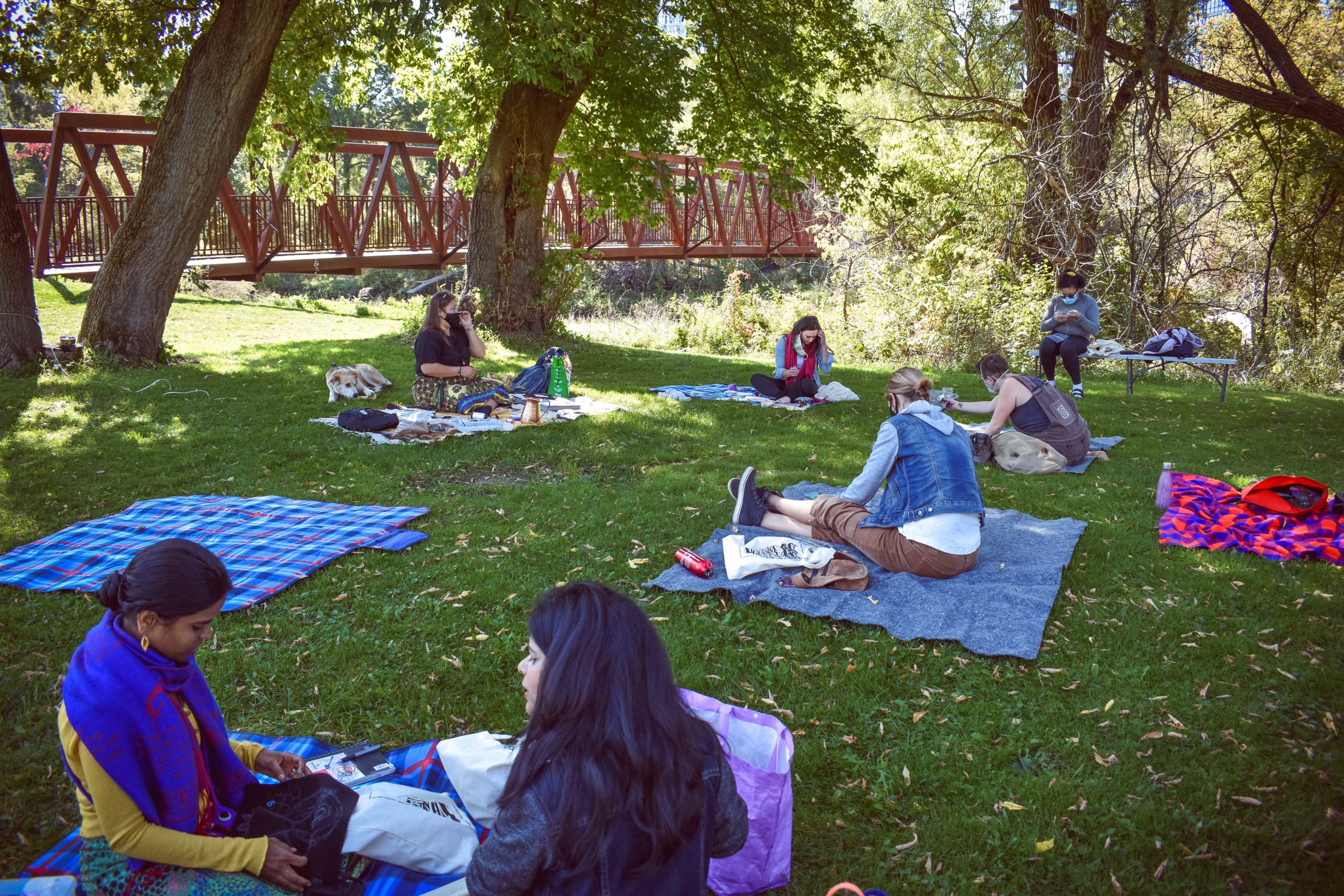 Photo of the Restoration of Relationship beading workshop, participants are sitting distant on their own picnic blankets on the grass. In the background there is foliages, trees and a bridge.