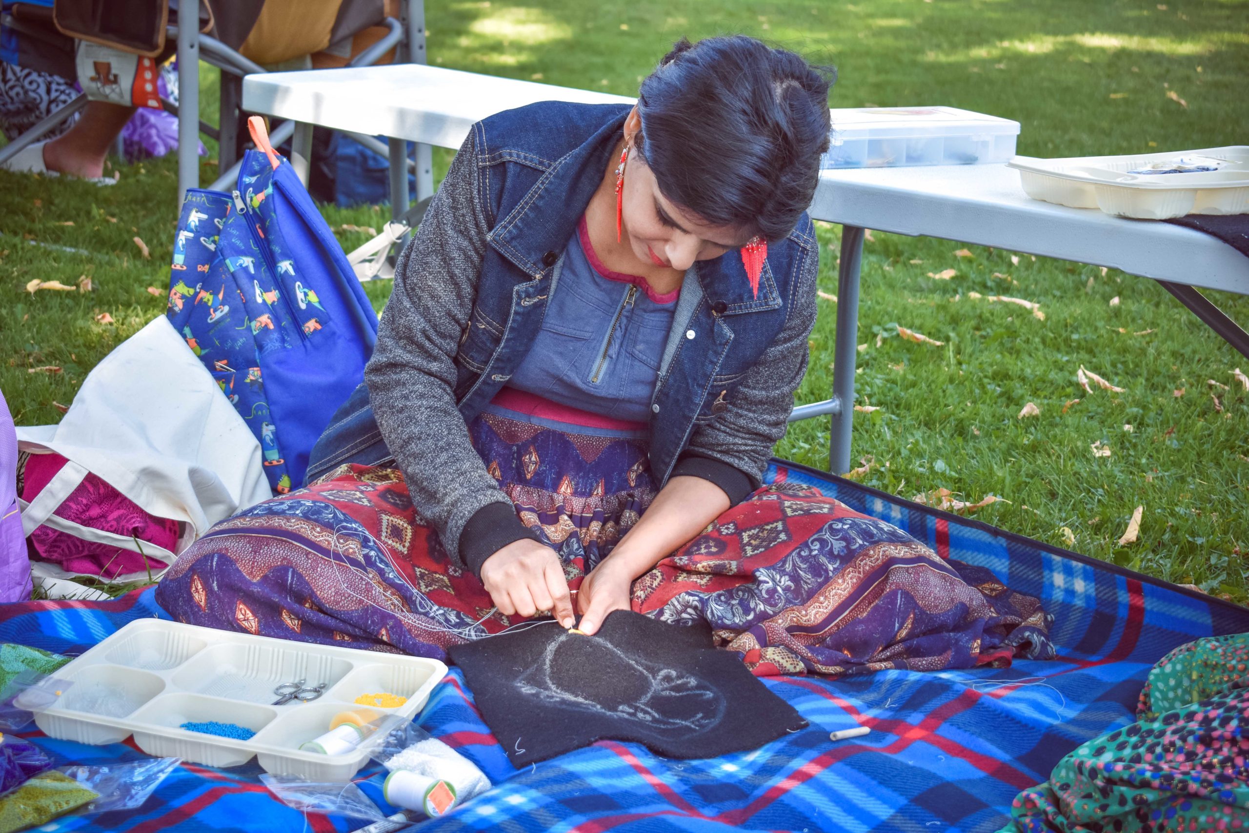 photo of a participant working on a beaded artwork, sitting on a blue picnic blanket. There are bags and trays with beads, a bench and table behind them