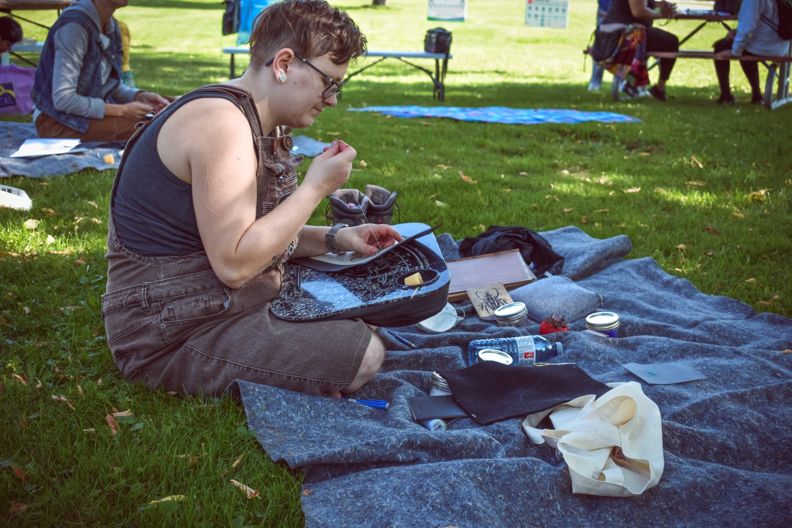 Photo of a person sitting on a grey blanket on the grass, working on their beaded artwork. There are items scattered around them, and other participants in the background sitting at picnic tables or blankets.