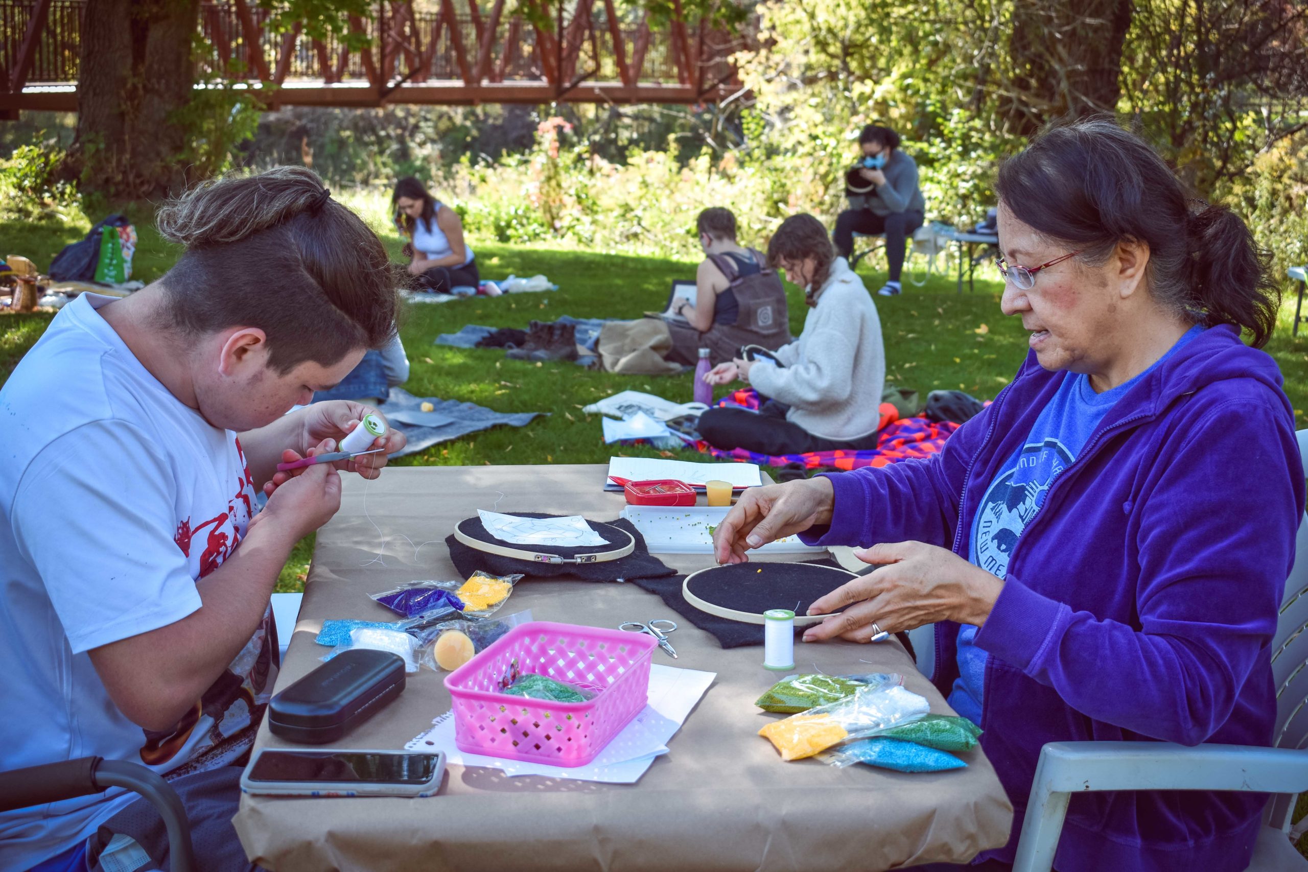 Photo of two people sitting at a picnic table working on their beaded artworks. The background has a grass, tables and other participants sitting on picnic blankets