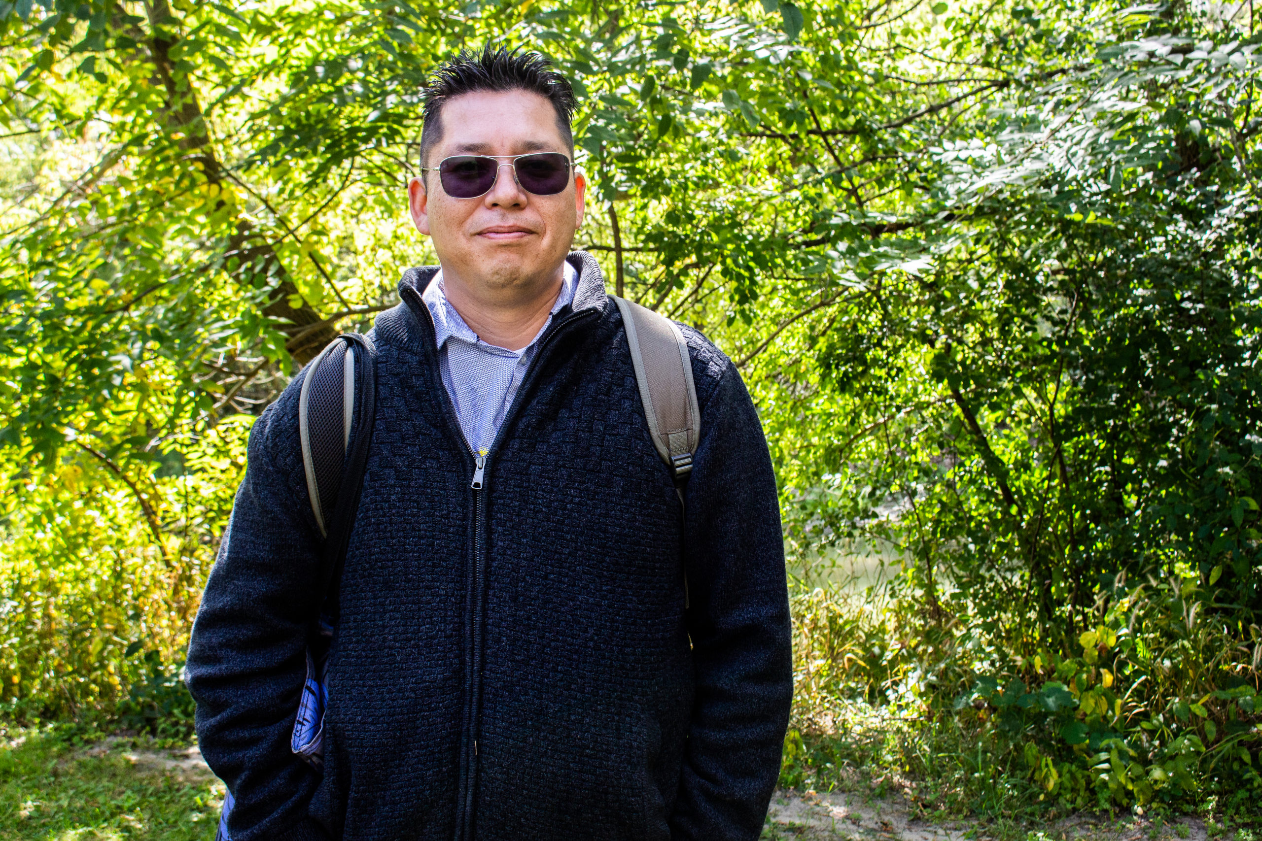 James Carpenter smiling at the camera and wearing dark sunglasses, a navy sweater and a backpack. James stands in front of a forest.