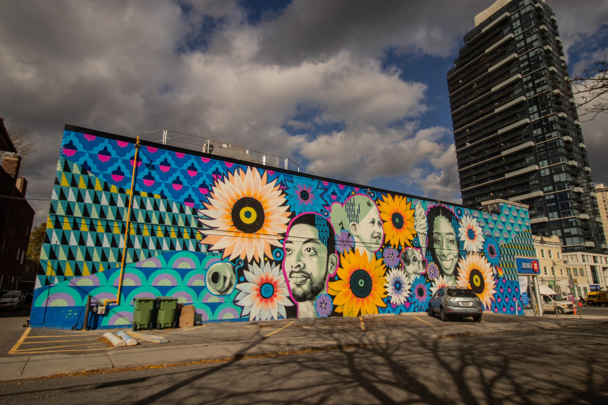 Photograph of Gosia's large-scale mural on the exterior wall of the BMO building, overlooking a parking lot. The mural features portraits of people in the community, animals, floral and geometric patterns.
