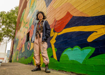 Photo of artist Wenting Li standing in front of her mural in Chinatown. It has three giant cranes intertwined with lotus flowers and roots, and carrying multiple human figures. It has colours of yellow, green, teal, red and pink.